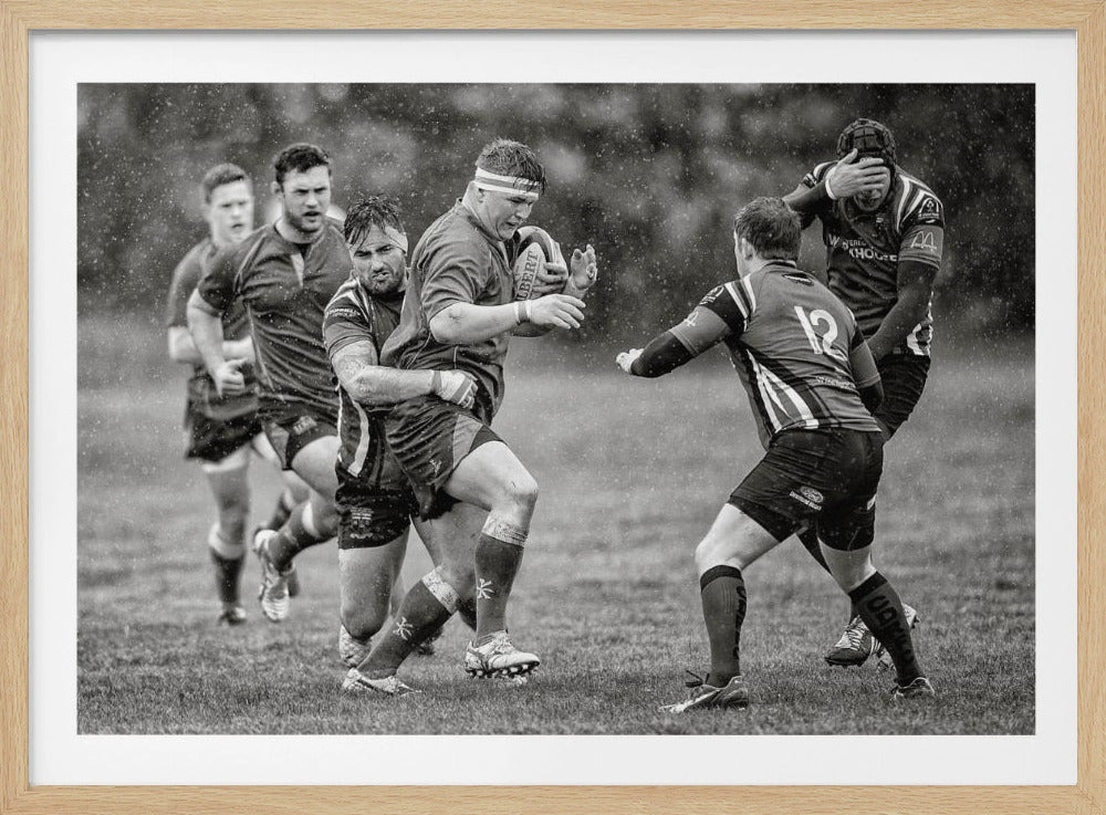 A dynamic black and white photograph of a rugby match being played in the rain. One player charges forward with the ball while an opponent wraps his arms around him in a tackle. Other players are in motion around them, capturing the intensity and physicality of the sport. Decor