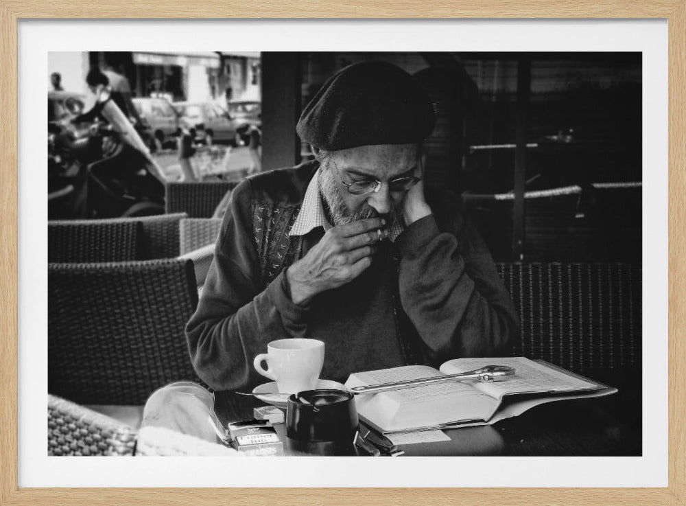 A framed, black and white photograph of an elderly man with a beard, glasses, and a beret, sitting at a cafe table. He is smoking a cigarette and looking down thoughtfully, with an open book, a coffee cup, and an ashtray on the table in front of him. Artwork