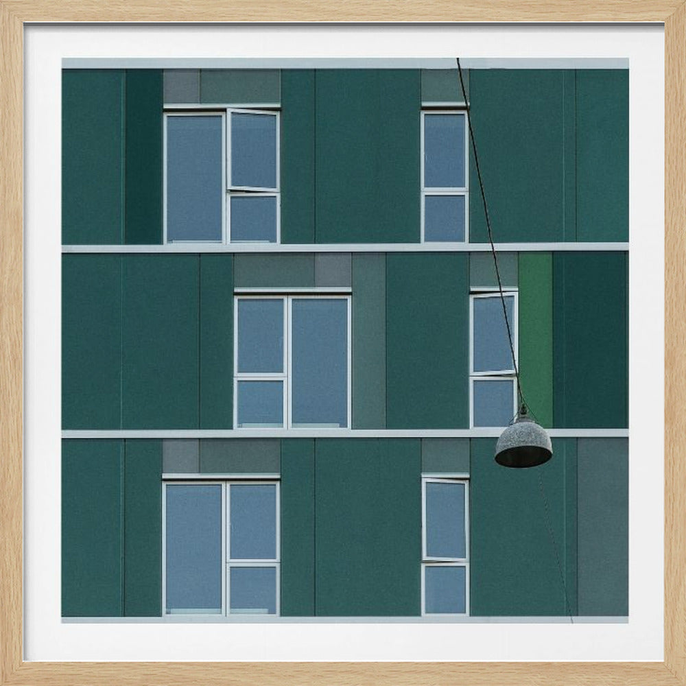 A framed photograph of a modern, dark green building facade with a grid of windows across three levels. A single industrial-style pendant lamp hangs in front of the building on the right side, interrupting the symmetrical pattern. The entire scene is enclosed by a light wood frame. Decor