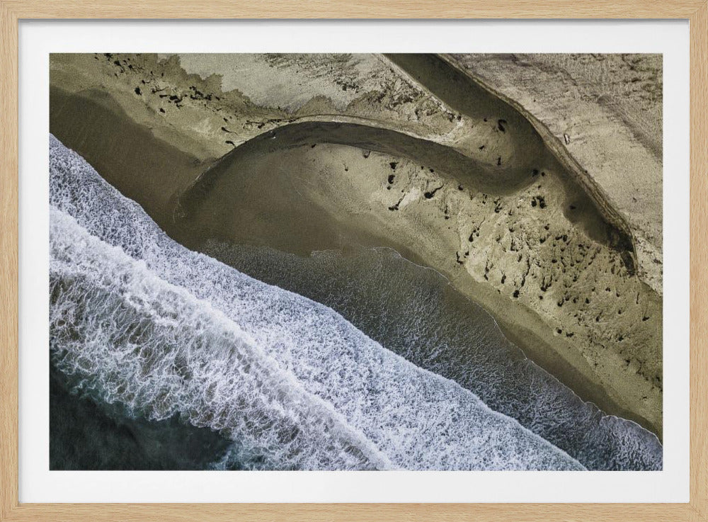 A framed aerial photograph of a coastline where white ocean waves crash diagonally against a sandy shore. A small river meanders through the beige sand, creating abstract, curved patterns as it flows towards the sea. Decor