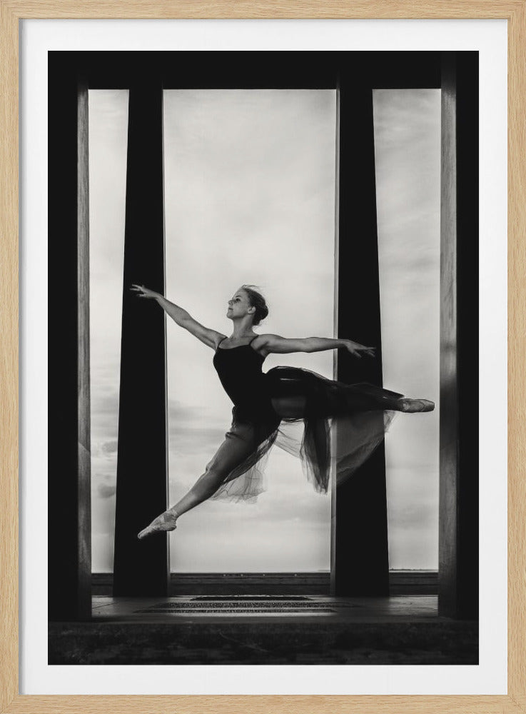 A powerful black and white photograph of a ballerina captured mid-leap. She is dressed in a dark leotard and a flowing, sheer skirt, with her arms and legs extended gracefully. The dancer is framed between large, dark vertical pillars against a bright, cloudy sky, creating a striking contrast and sense of movement. Print