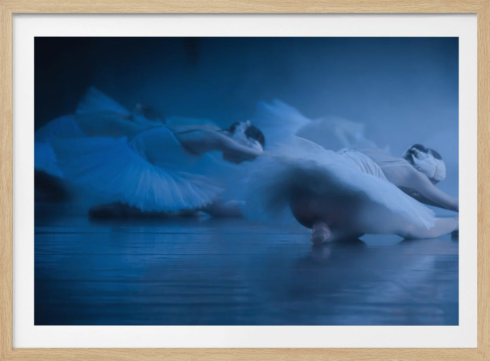 A low-angle, motion-blurred photograph of ballerinas in white tutus performing on a stage. The scene is bathed in a dramatic blue light, emphasizing the graceful movement of the dancers. The image is presented within a silver frame. Artwork