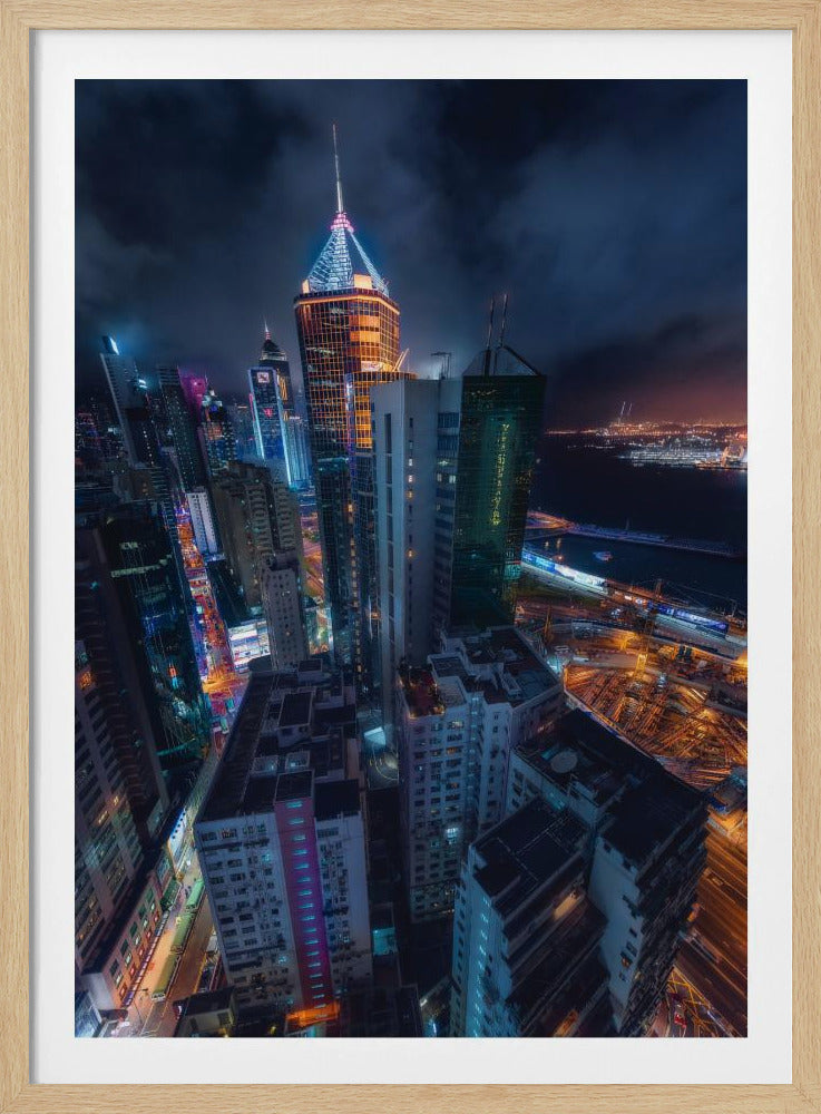 A dramatic high-angle nighttime photograph of a sprawling modern cityscape, with towering skyscrapers illuminated in vibrant blue, orange, and purple neon lights under a dark, cloudy sky. Below, streets are aglow with traffic, and in the distance, a brightly lit harbor meets the dark water. Decor