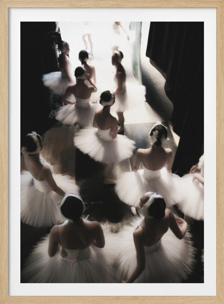 A high-angle, soft-focus photograph capturing a group of ballerinas in white tutus from behind as they wait in the dark wings of a stage, with a bright light illuminating the area ahead of them. Artwork
