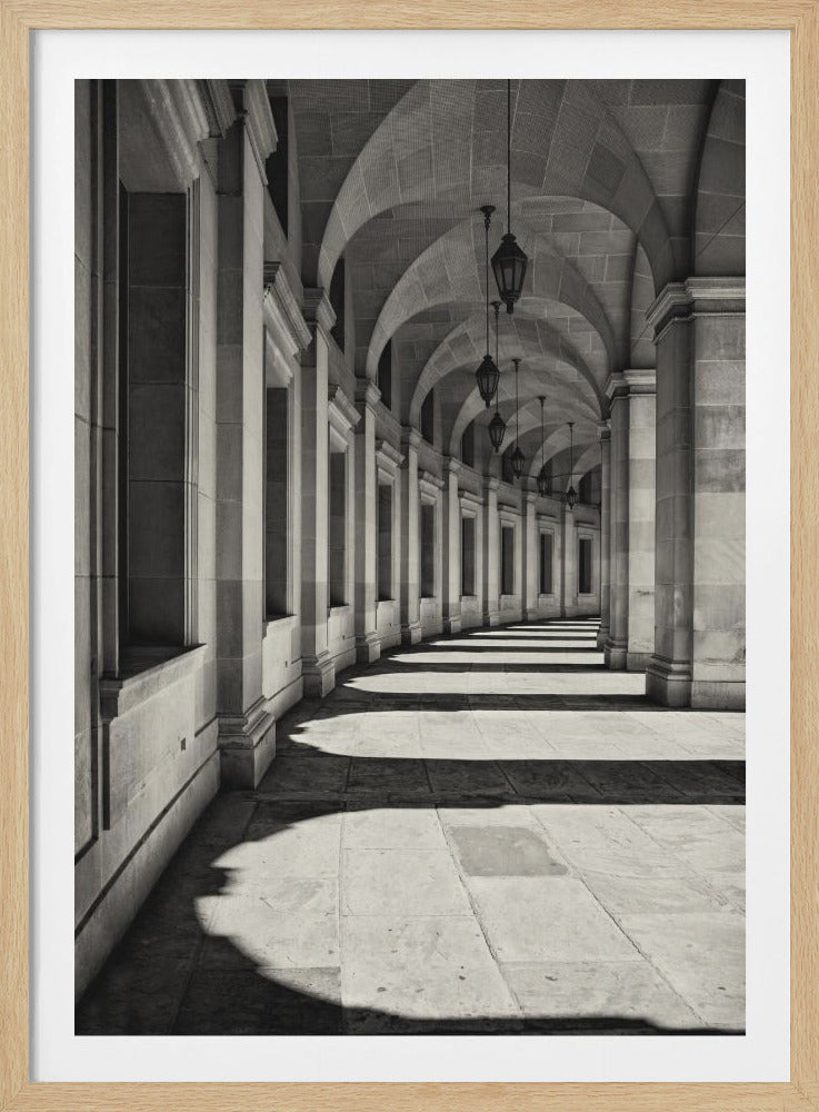 A black and white photograph of a long, curving stone colonnade. Sunlight creates a dramatic play of light and shadow, casting long shadows from the pillars across the floor. Ornate lanterns hang from the vaulted, arched ceiling, leading the eye down the corridor. Wall Art
