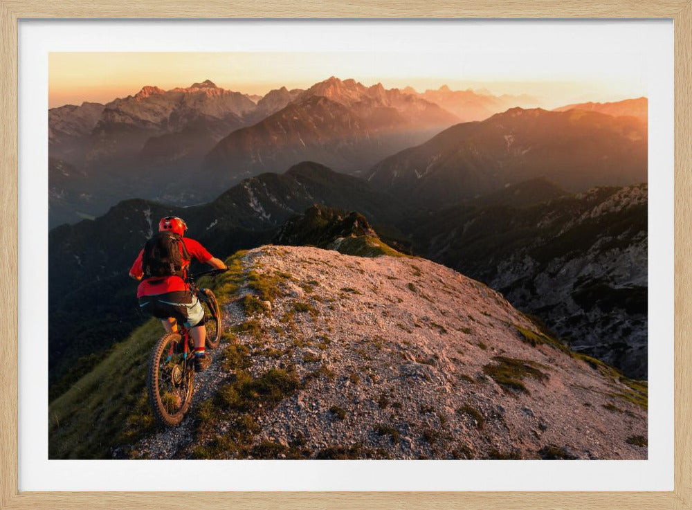 A mountain biker wearing a red shirt and helmet rides along a narrow, rocky mountain ridge at sunset. The background features a vast landscape of layered mountain ranges glowing in the warm, golden light of the setting sun. Poster