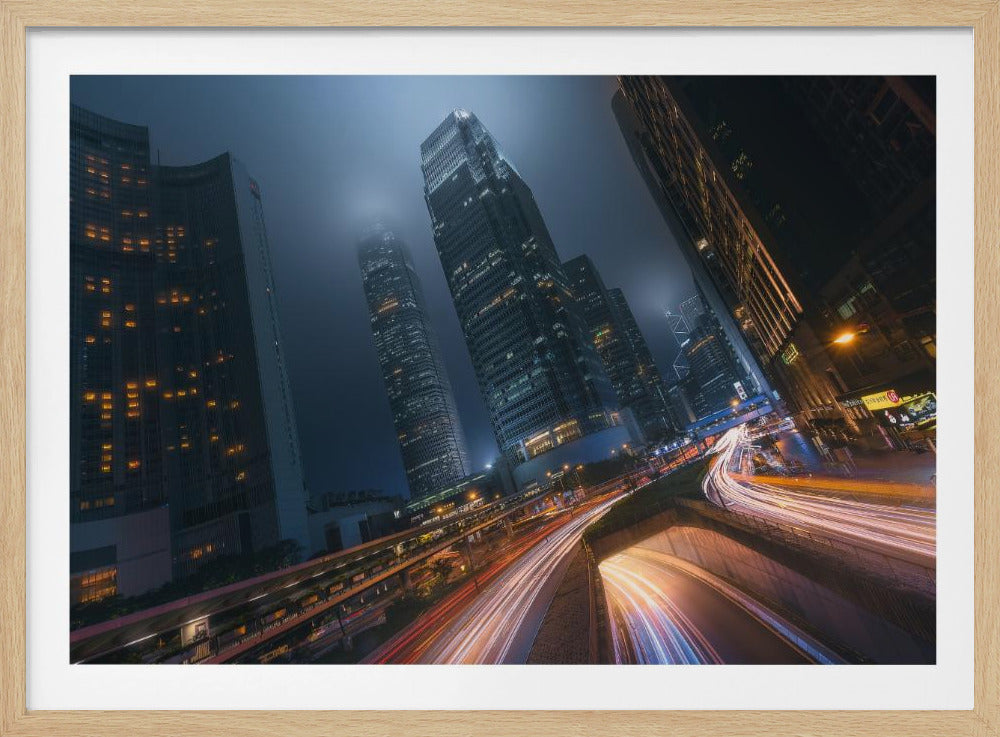 A framed, low-angle photograph capturing a bustling city at night, with towering skyscrapers shrouded in fog. Long-exposure creates vibrant streaks of light from traffic on a multi-level highway, illuminating the dark, modern urban landscape. Wall Art