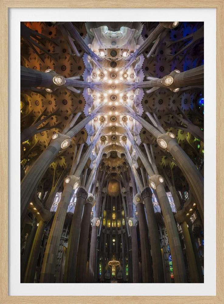 A low-angle, symmetrical photograph looking up at the incredibly ornate and complex vaulted ceiling of the Sagrada Familia Basilica. The massive stone columns branch out like trees, supporting the intricately detailed ceiling illuminated by warm, glowing lights and splashes of color from unseen stained glass windows. Poster