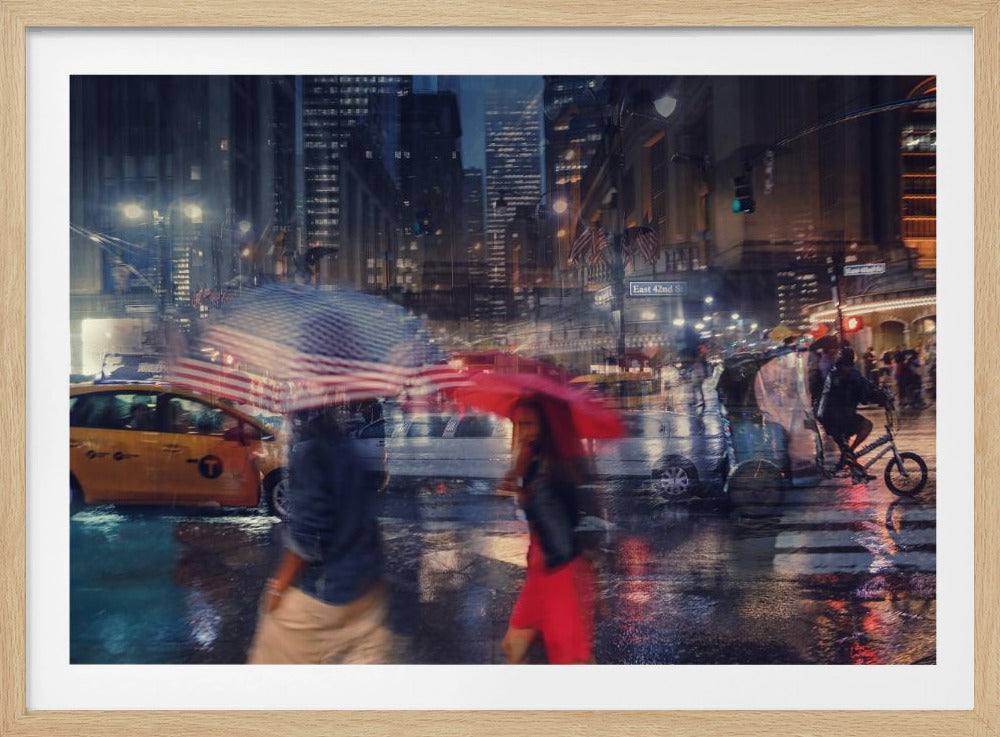 An artistic motion-blur photograph of a rainy night on East 42nd Street in New York City. Pedestrians with umbrellas, one with an American flag design and another bright red, cross the wet street as city lights and a yellow taxi reflect on the pavement, creating a vibrant, impressionistic scene within a silver frame. Decor