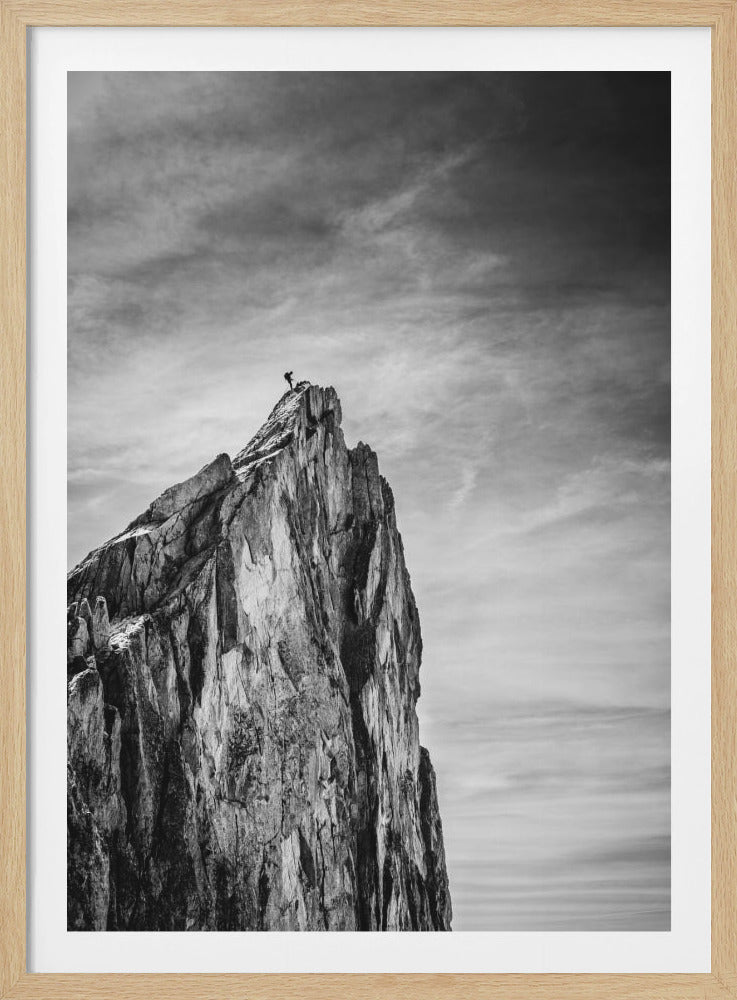 A dramatic black and white photo of a lone climber silhouetted on the very summit of a craggy, imposing mountain peak, set against a vast, cloudy sky. Artwork