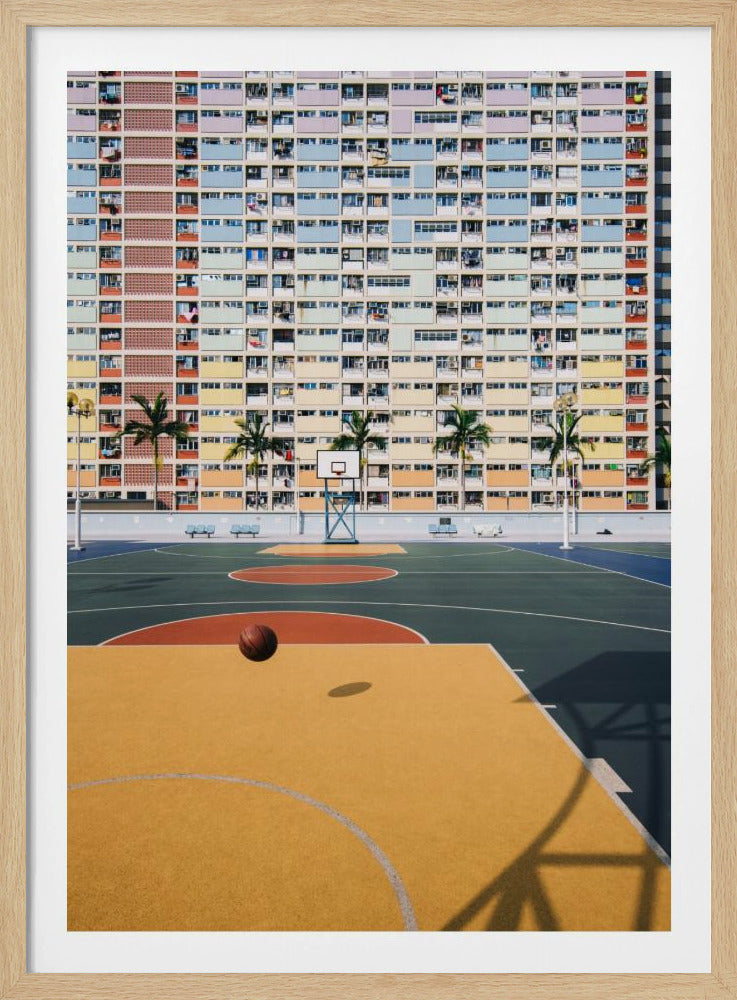 A vibrant photograph of a basketball court with a yellow and green floor, a single basketball is mid-bounce in the foreground. In the background stands the massive, symmetrical Choi Hung Estate apartment building in Hong Kong, famous for its facade of pastel rainbow colors. Palm trees line the space between the court and the building. Print
