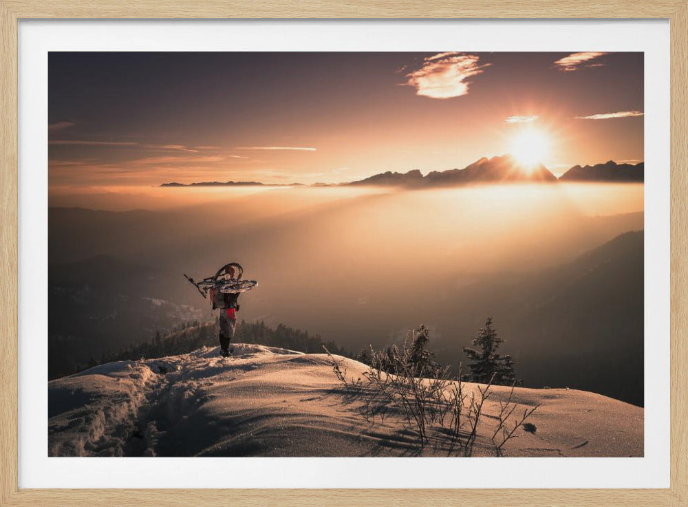 A lone person stands on a snowy mountain peak at sunrise, carrying a bicycle over their shoulder. They look out over a valley filled with a sea of clouds, with distant mountain ranges silhouetted against the bright, golden sun. Artwork