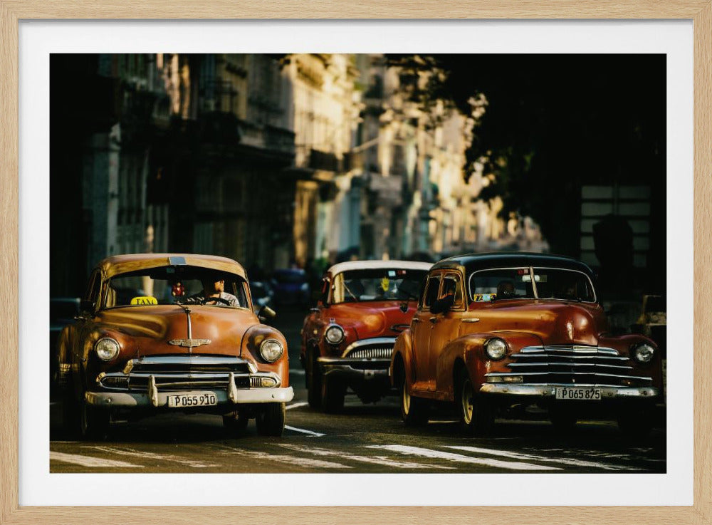 Three vintage American cars drive down a sunlit city street. The foreground features an orange taxi, with a red car and another orange vehicle behind it, all under the warm glow of late afternoon light casting long shadows. Print