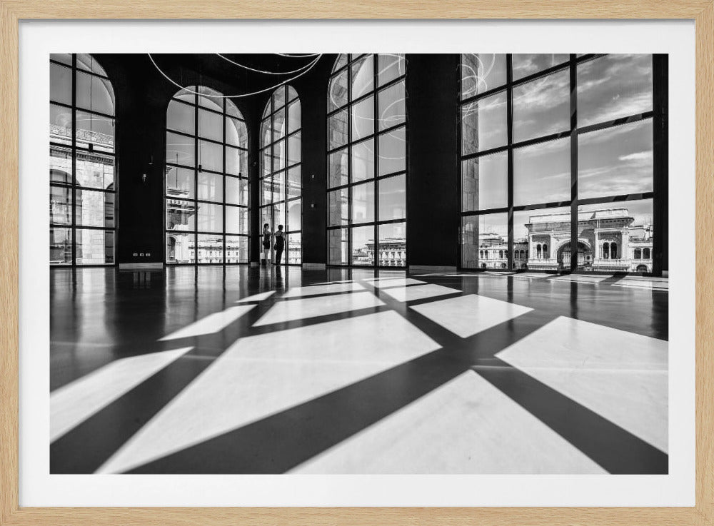 A striking black and white photograph of a spacious, modern hall with a polished floor. Dramatic shadows from large, arched windows are cast across the floor. Through the windows, a view of a classical European city with a triumphal arch can be seen. Print