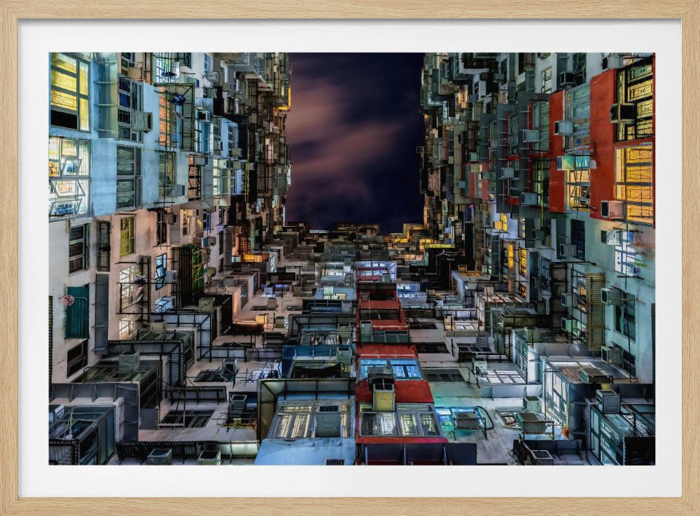 A low-angle nighttime view looking up from a courtyard surrounded by extremely dense, stacked apartment buildings with many windows lit in yellow, blue, and white, revealing a small patch of dark, cloudy sky. The image is presented within a brushed silver frame. Artwork