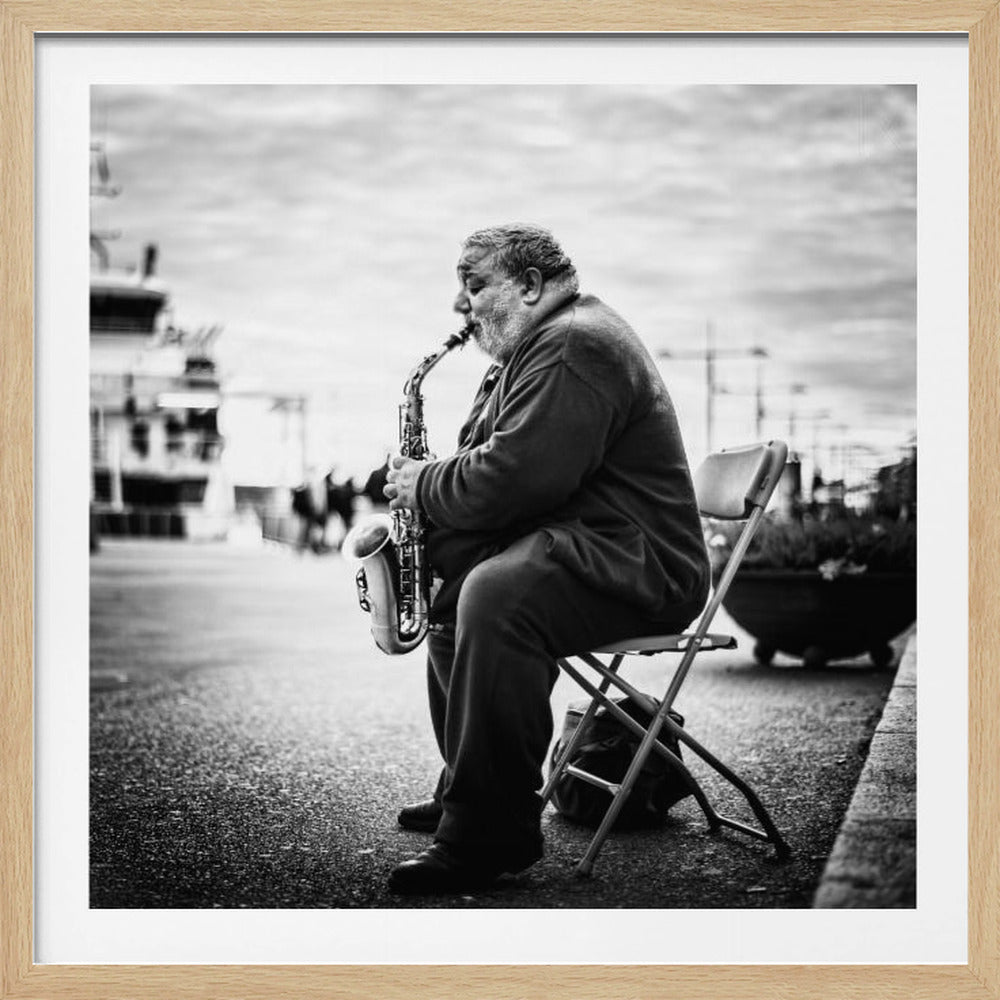 A black and white framed photograph of an older, heavyset man sitting in profile on a folding chair on the street, playing a saxophone with his eyes closed. The background is a blurry urban or harbor scene. Artwork