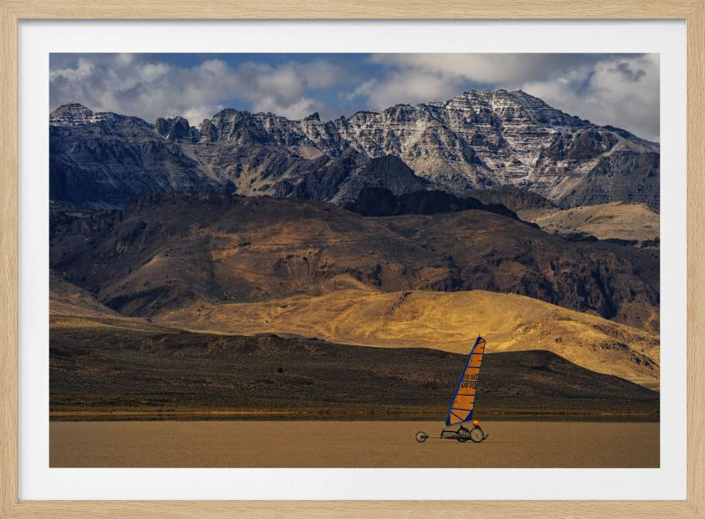 A framed photograph of a land sailor with a bright orange and blue sail on a vast, flat desert floor. In the background, majestic snow-dusted mountains rise against a cloudy blue sky. Artwork