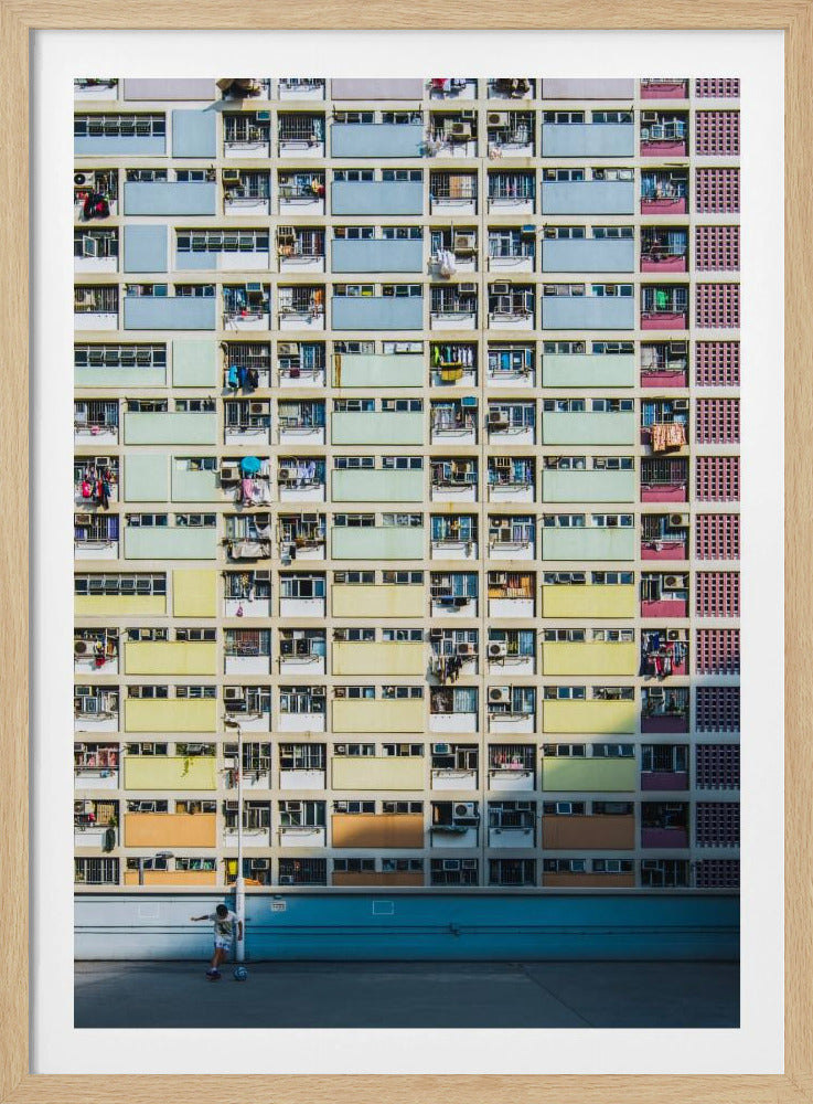 A symmetrical, full-frame shot of a massive apartment building with a repeating grid pattern of windows and balconies. The building is painted in horizontal bands of pastel colors, starting with blue at the top, then green, yellow, and orange. A single boy plays with a soccer ball on the ground level in front of a blue wall. Wall Art