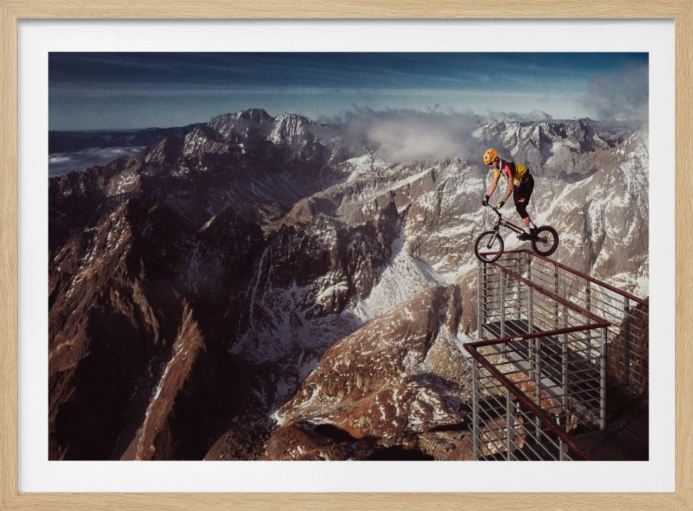 A trials cyclist wearing a yellow helmet balances precariously on the railing of a metal walkway high in the mountains, with a dramatic background of snow-dusted peaks and a blue sky. Poster