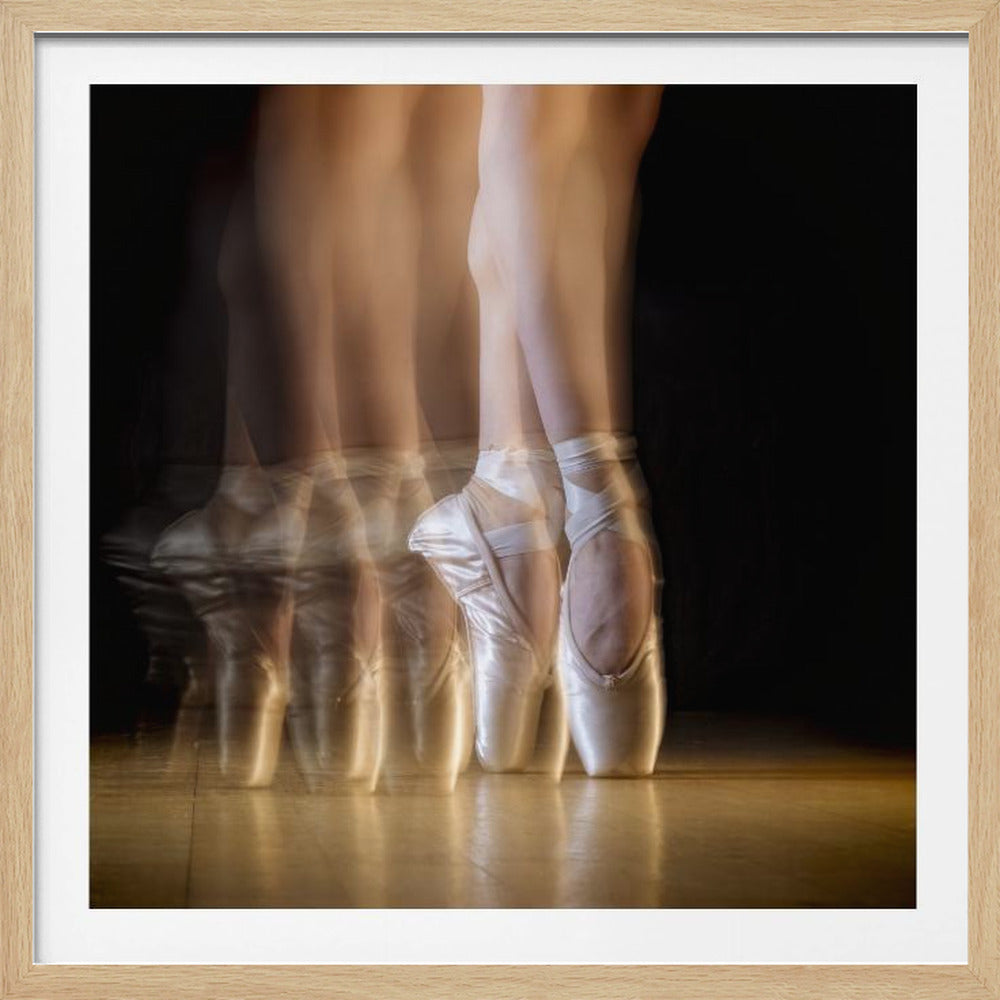 A long-exposure photograph showing the lower legs and feet of a ballerina in pointe shoes, capturing the motion of a dance step. Multiple blurred images of the feet transition from left to right, ending in a sharp, clear image of the final pose. The scene is set on a reflective wooden floor against a solid black background, encased in a light wood frame. Artwork