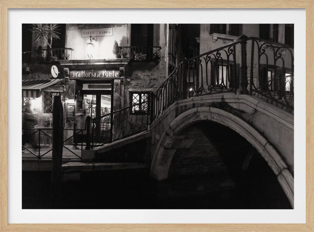 A black and white photograph of a Venetian canal at night. On the right, a stone bridge with an ornate iron railing arches over the dark water. On the left, the entrance to a restaurant named 'Trattoria al Ponte' is illuminated, casting a warm glow on the weathered building facade. Print