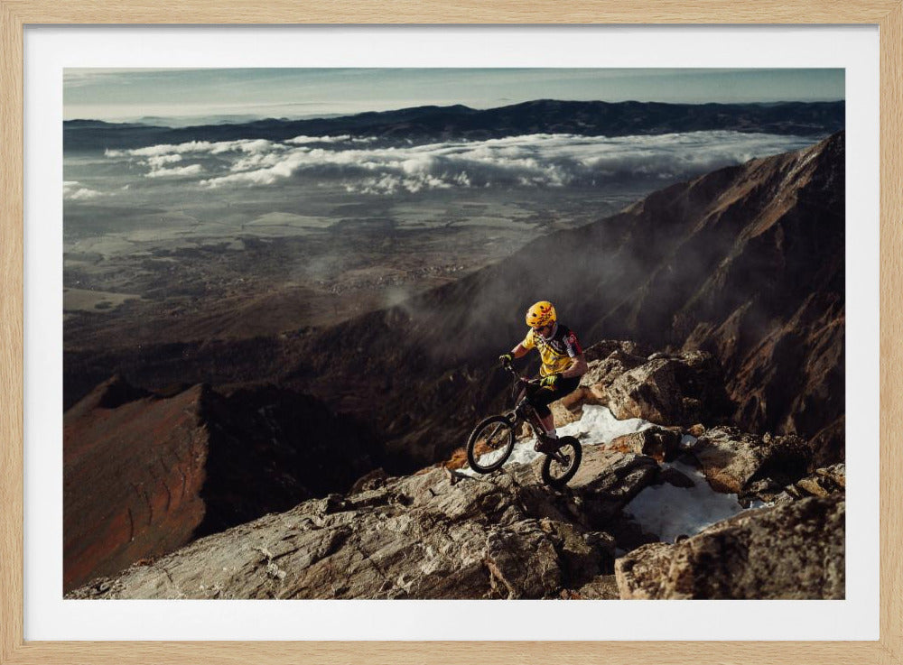 A mountain biker in a yellow helmet and jersey navigates a treacherous, rocky mountain ridge. Below, a vast valley is filled with a sea of clouds, with distant mountain ranges visible on the horizon. Print