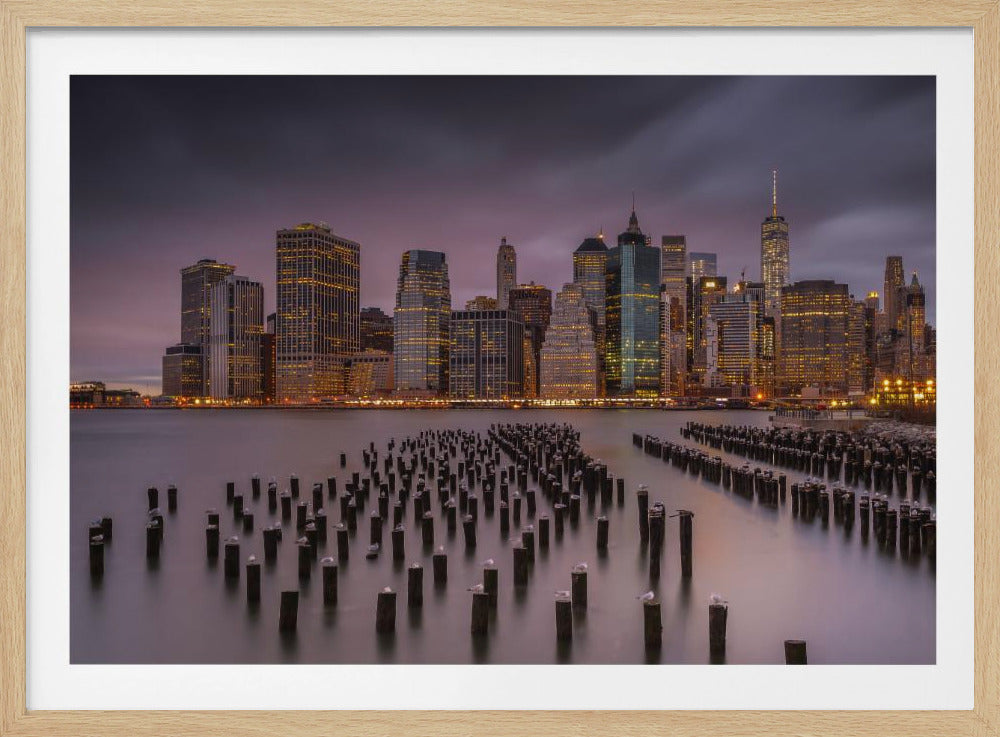 A long exposure photograph of the Lower Manhattan skyline at dusk, viewed from across the water. The city buildings are illuminated with warm yellow lights against a dark purple, cloudy sky. In the foreground, hundreds of old wooden pier pilings stick out of the calm water, with several white seagulls perched on top. Decor