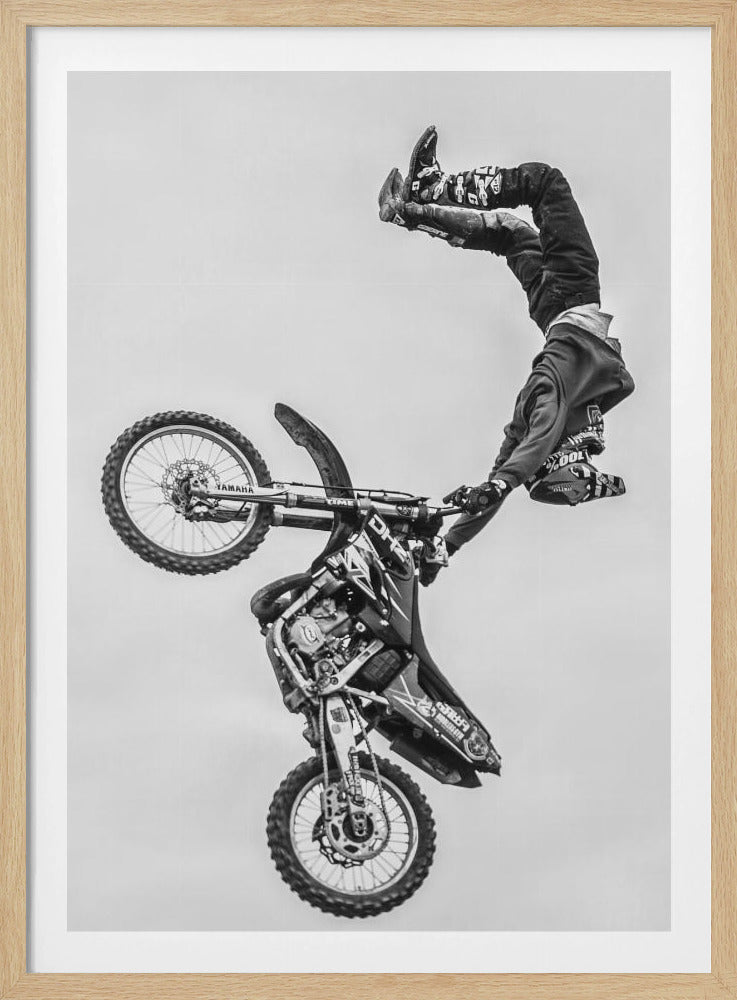 A dynamic black and white action shot of a freestyle motocross rider performing a handstand on the handlebars of their dirt bike mid-air. The bike is nearly vertical against a plain white sky, capturing a moment of extreme athleticism and balance. Poster