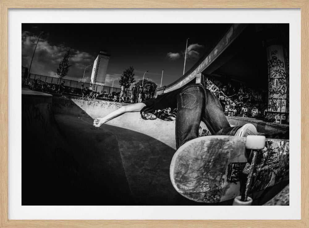 A dynamic, wide-angle black and white photograph of a skateboarder at a skatepark. The skater is bent backwards over the curved edge of a concrete bowl, with one hand touching the surface. Their skateboard is propped up in the foreground, and the background is filled with graffiti-covered walls, spectators, and a tall building against a dark, cloudy sky. Artwork