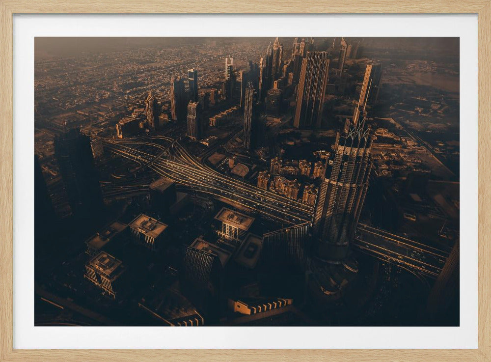 An aerial view of a sprawling modern city at dusk, with the warm glow of sunset casting long shadows over skyscrapers and illuminating a complex network of highways. The photograph has a warm, sepia-like tone and is presented within a silver-colored frame. Decor