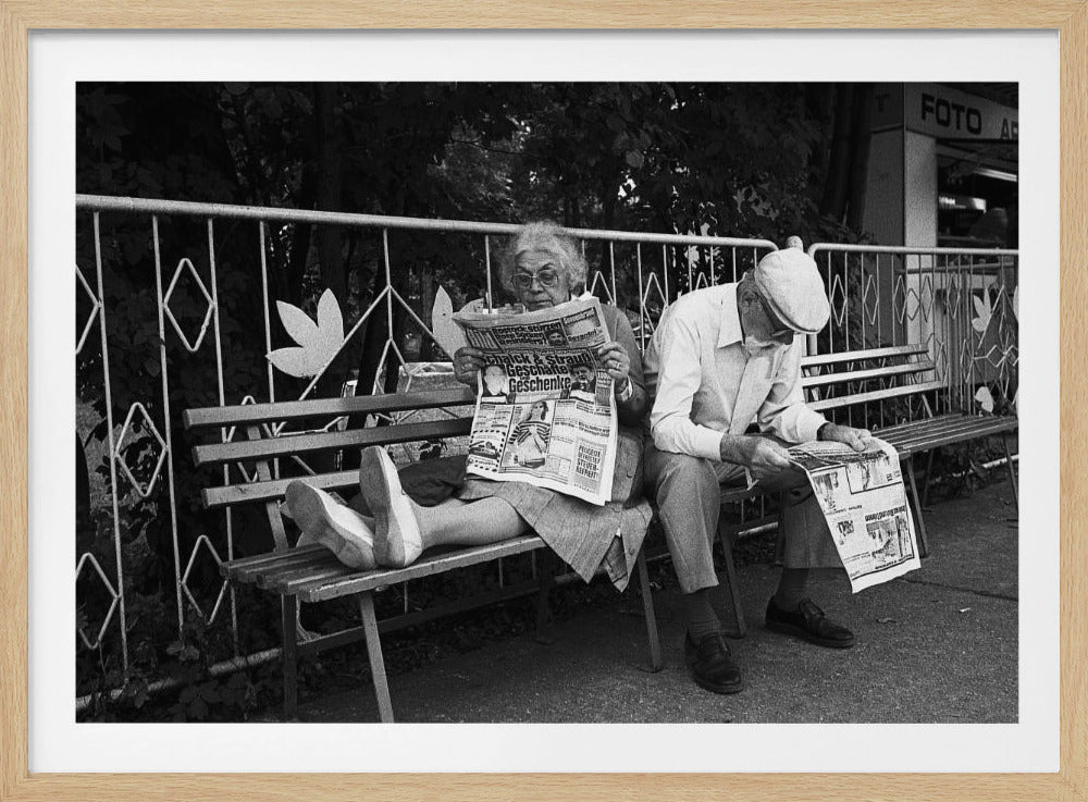 A framed black and white photograph of an elderly couple sitting on a park bench, both deeply absorbed in reading their newspapers. The woman on the left has her feet propped up on the bench, while the man on the right sits next to her, looking down at his paper. Poster
