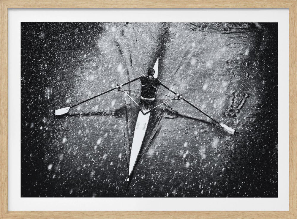 A dramatic black and white overhead photograph of a single rower in a narrow scull, captured mid-stroke as they cut through dark water during a heavy snowfall, creating a high-contrast and atmospheric scene. Decor