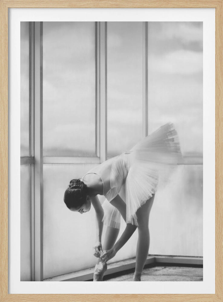 A black and white photograph of a ballerina in a white tutu, bending over to tie her pointe shoe in front of a large, bright window. The motion of her tutu is captured in a graceful blur. Print