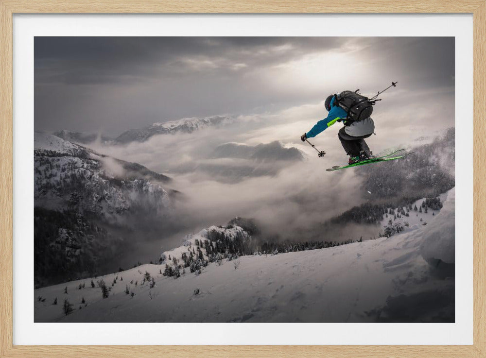 A skier wearing a blue jacket and a backpack is captured mid-air, jumping off a snowy mountain ridge against a dramatic backdrop of cloud-filled valleys and distant snow-capped peaks. Poster