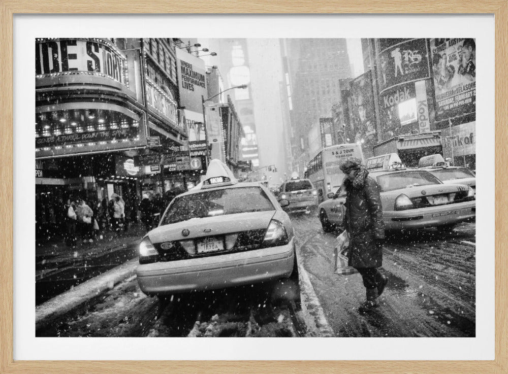 A black and white photograph of a bustling New York City street during a snowstorm. A person in a heavy winter coat crosses the street in front of a line of yellow cabs, with the bright lights of Times Square billboards glowing softly in the background through the falling snow. Print