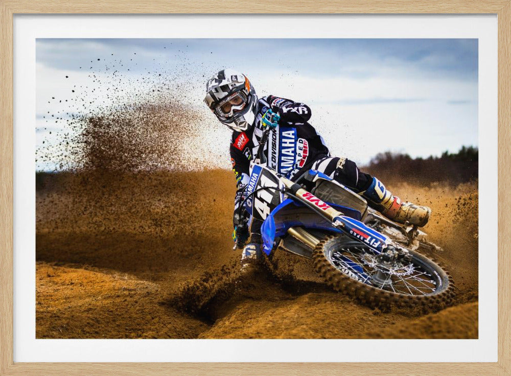 A motocross rider in full gear leans sharply into a turn on a dirt track, kicking up a large spray of mud and dirt from their blue and white Yamaha motorcycle. Artwork
