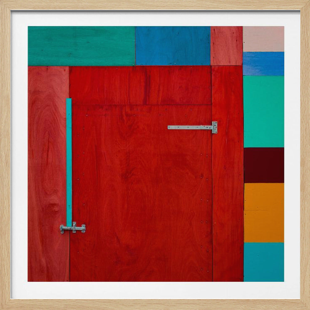 A framed, square abstract artwork featuring a large, vibrant red wooden door with silver metal latches. The door is surrounded by a collage of other wooden panels painted in solid blocks of color, including teal, blue, orange, and maroon, creating a geometric, color-block composition. Artwork
