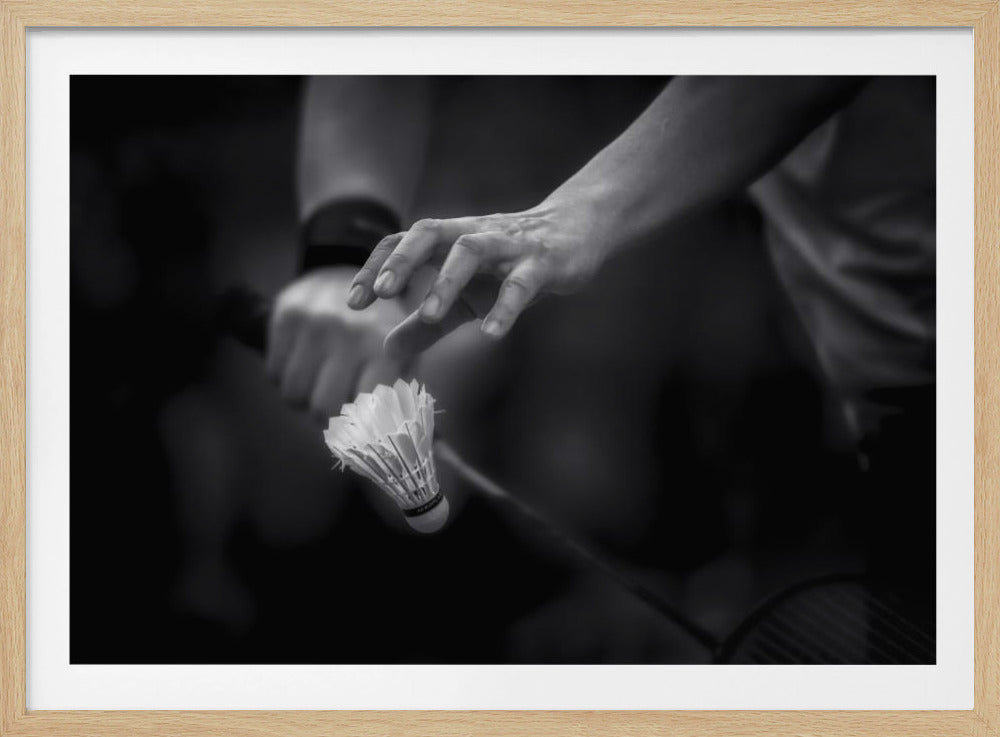 A dramatic black and white close-up photograph of a badminton player preparing to serve. The focus is on the player's hands, with one hand delicately positioned above a shuttlecock which is balanced on the strings of a racquet. Artwork