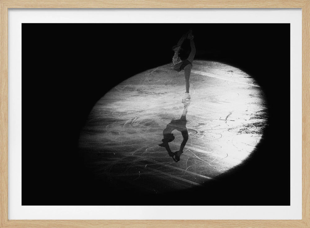 A dramatic black and white framed photograph of a female figure skater performing on ice. She is illuminated by a single spotlight, holding a graceful standing split pose, with her reflection visible on the ice below. Poster
