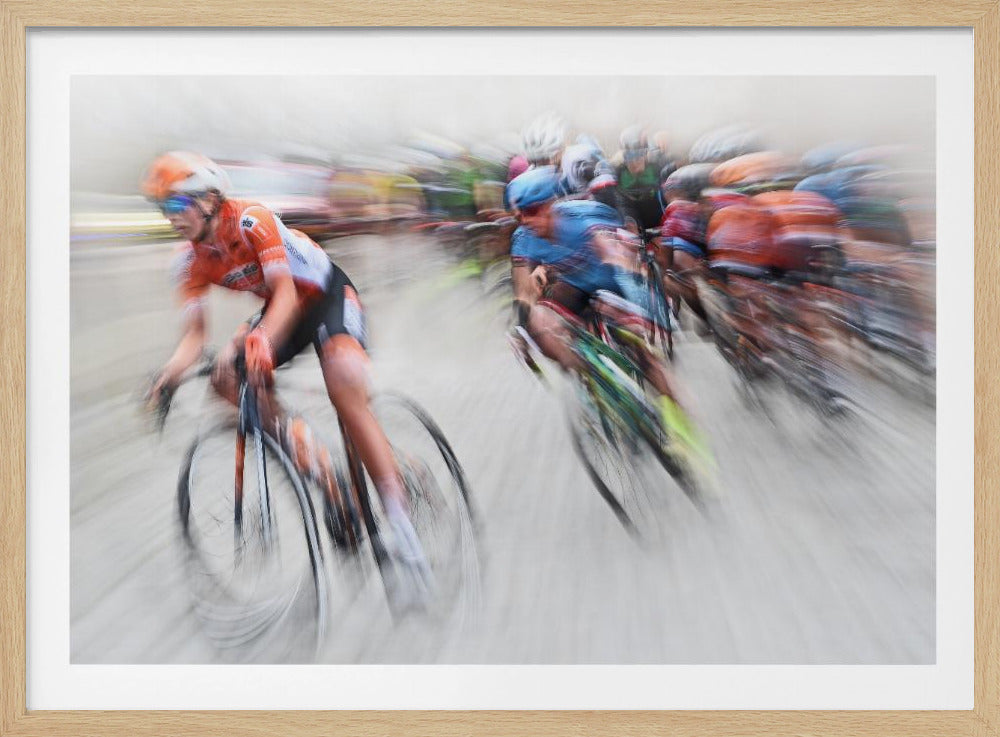 An action photograph of a professional cycling race captured with a prominent motion blur effect that conveys a strong sense of speed. A cyclist in an orange and black uniform is in the foreground, leaning into a turn, while a group of other riders in blue and multi-colored jerseys follows closely behind, their forms streaked and blurred. The entire scene is encased in a brushed silver frame. Poster
