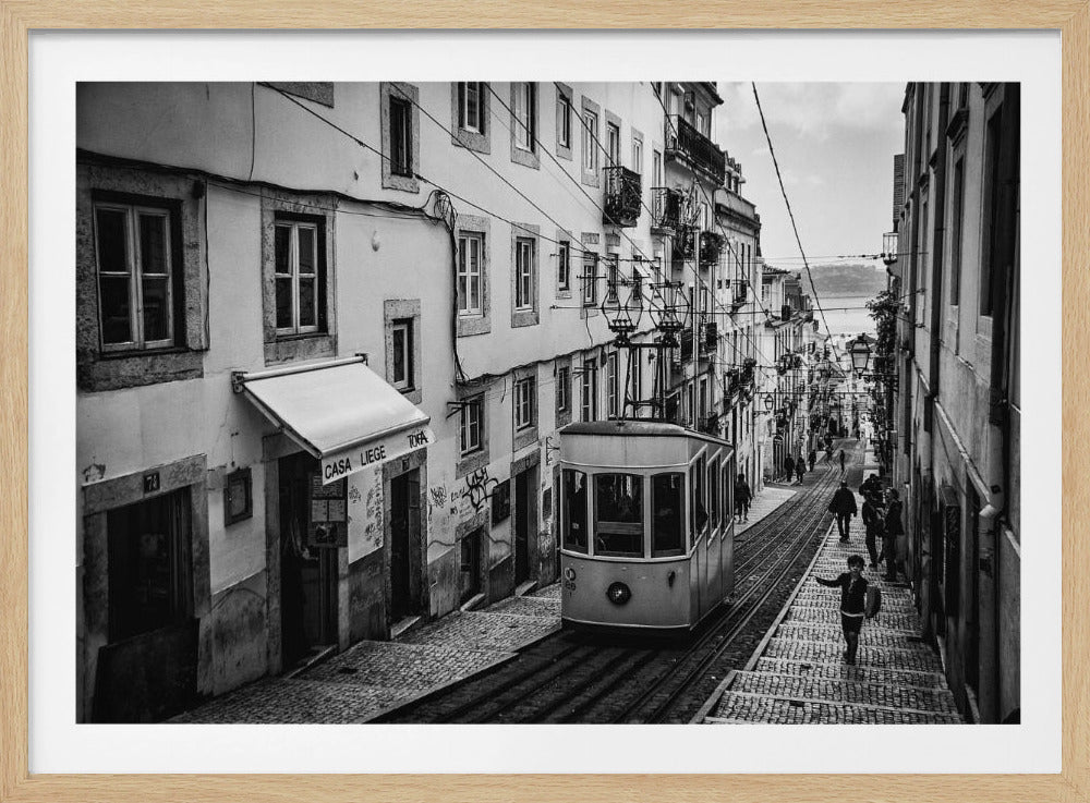A black and white photograph of a vintage tram on a steep, narrow cobblestone street in an old European city. The street is lined with historic buildings and people are walking alongside the tracks. In the distance, a body of water is visible. Poster