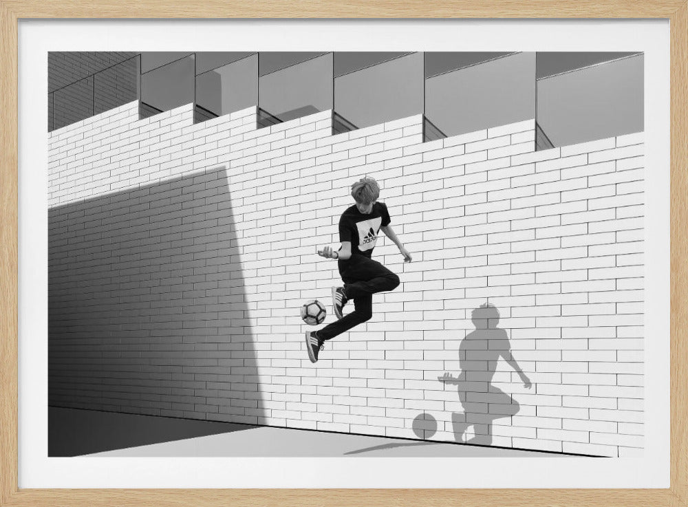 A framed, black and white photograph of a young person in mid-air, skillfully juggling a soccer ball with their foot against a clean, white brick wall. The person's shadow is cast sharply on the wall, mirroring the action, under a modern building with angled windows. Wall Art