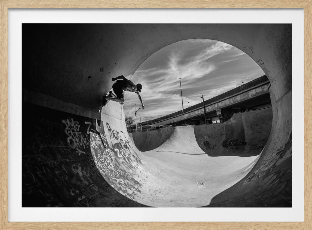 A dynamic black and white action shot of a skateboarder mid-trick on the edge of a concrete ramp in a graffiti-covered skatepark, framed by the curve of a large pipe. Print