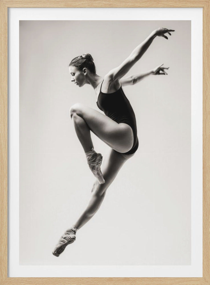 A black and white studio photograph of a graceful ballerina captured mid-leap. She wears a dark leotard and pointe shoes, her body contorted in a dynamic pose against a plain, light background, framed in black. Wall Art