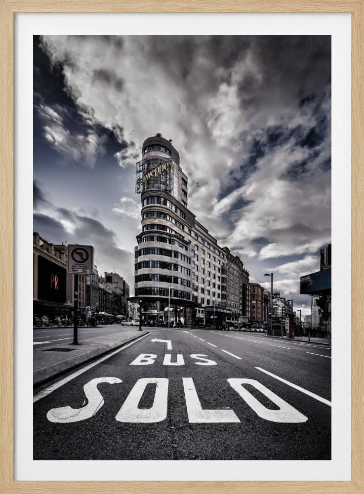 A dramatic, low-angle, black and white photograph of the iconic Edificio Carrión in Madrid. The perspective is from the middle of a street, with the words 'SOLO BUS' painted in large white letters on the asphalt in the foreground. The building, famous for its large 'Schweppes' sign, stands tall against a moody, cloud-filled sky. Artwork