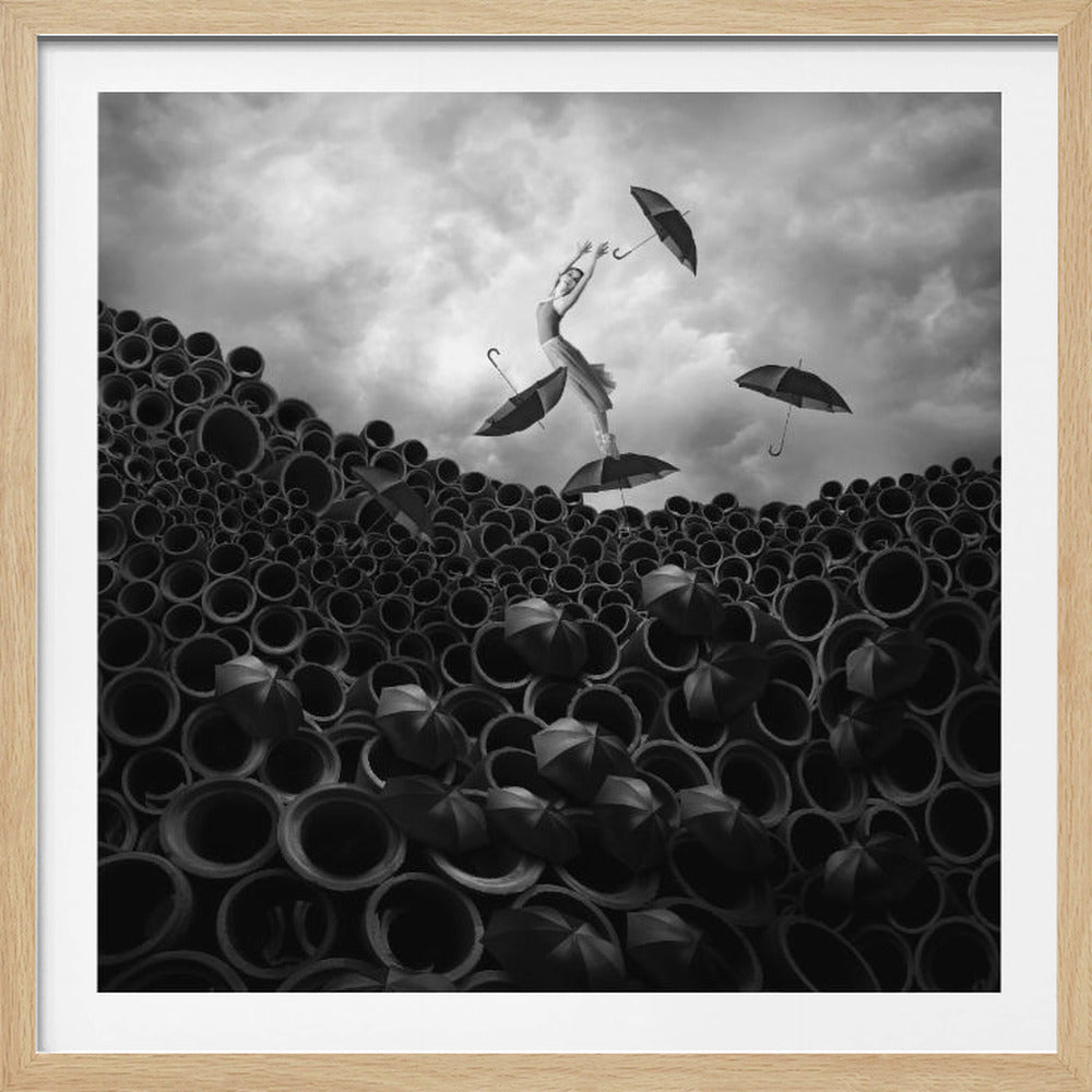 A surreal black and white photograph of a woman balancing on an umbrella atop a large pile of industrial pipes. Several other umbrellas are floating in the air around her and sitting in the openings of the pipes below, all set against a dramatic, cloudy sky and enclosed in a light wood frame. Poster