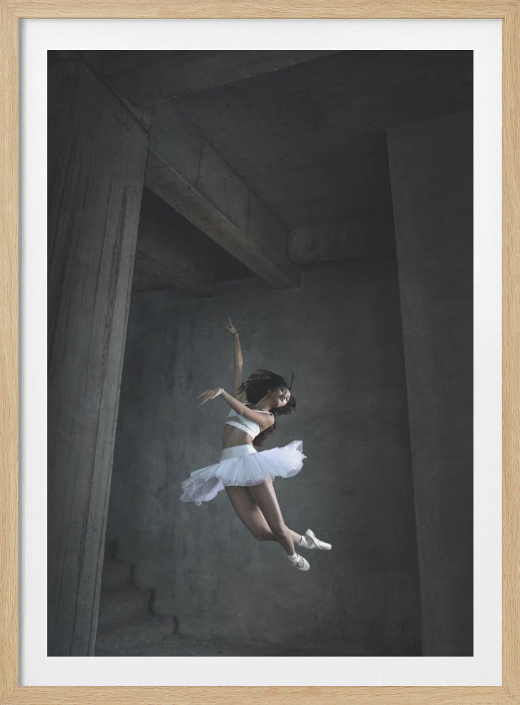 A graceful ballerina in a white tutu is captured mid-air, her body arched in a dynamic dance pose. She is set against a stark, moody background of gray concrete pillars and beams in an industrial-style space. Print