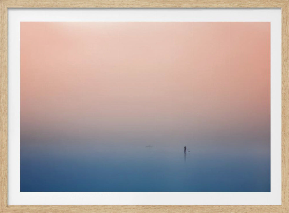 A minimalist framed photograph of a solitary person on a stand-up paddleboard, silhouetted against a vast, calm expanse of water under a hazy, gradient sky that shifts from soft pink to deep blue. Wall Art