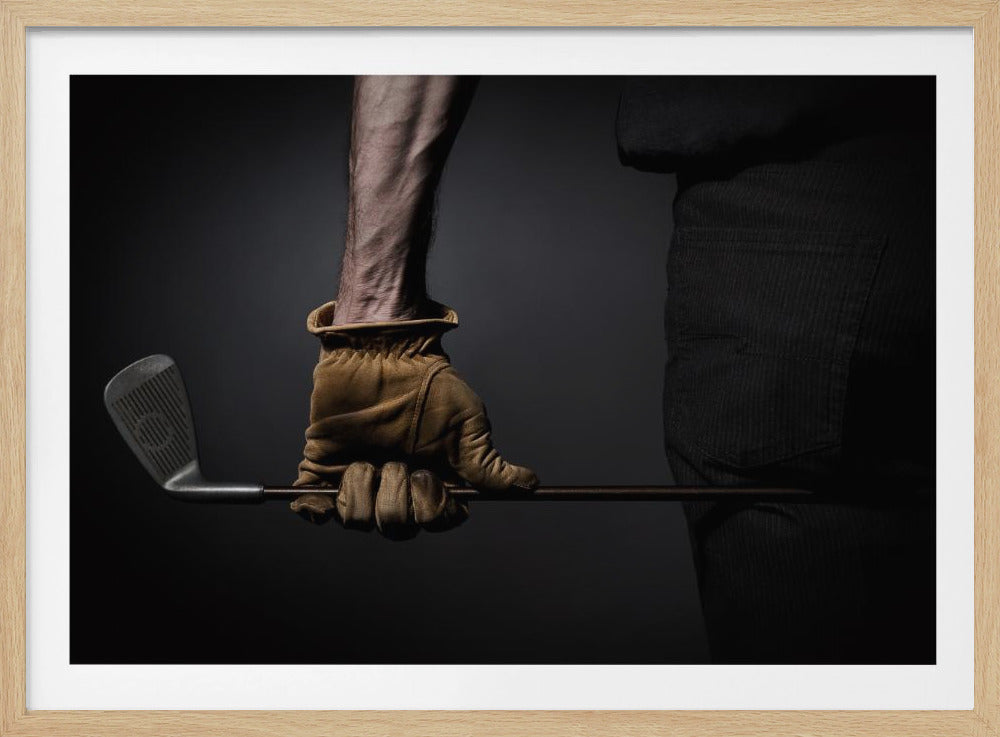 A dramatic close-up of a person's muscular forearm and hand wearing a tan leather glove, holding a golf iron horizontally against a dark background, framed in silver. Wall Art