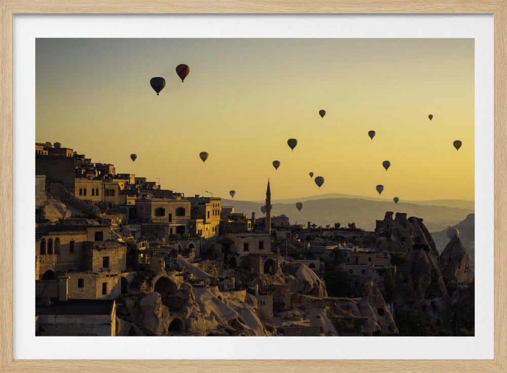 A wide-angle photograph of numerous hot air balloons floating over the ancient town of Cappadocia, Turkey, during a golden sunrise. The sky is a warm yellow gradient, and the stone buildings and unique rock formations of the town are cast in shadow and morning light in the foreground. Wall Art